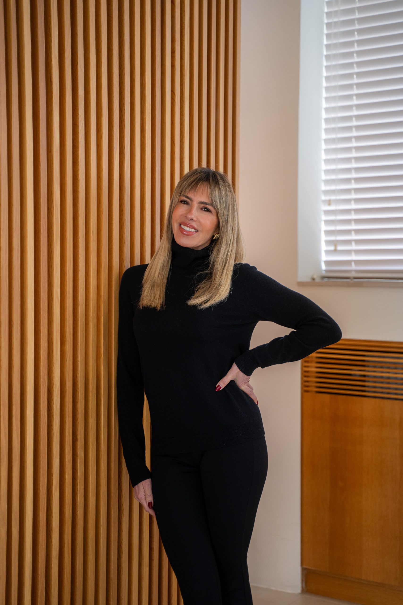 Esther Sarphatie smiling while standing beside a wooden slat wall, wearing a black outfit.
